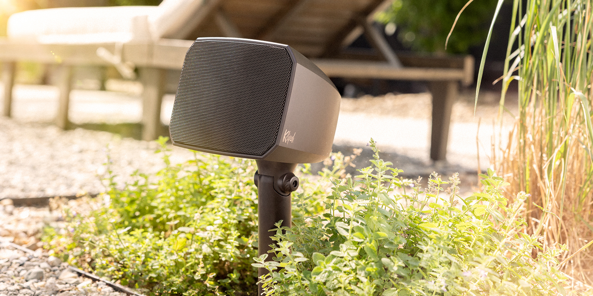 Klipsch Courtyard speaker planted in greenery on outdoor courtyard patio in sunlight